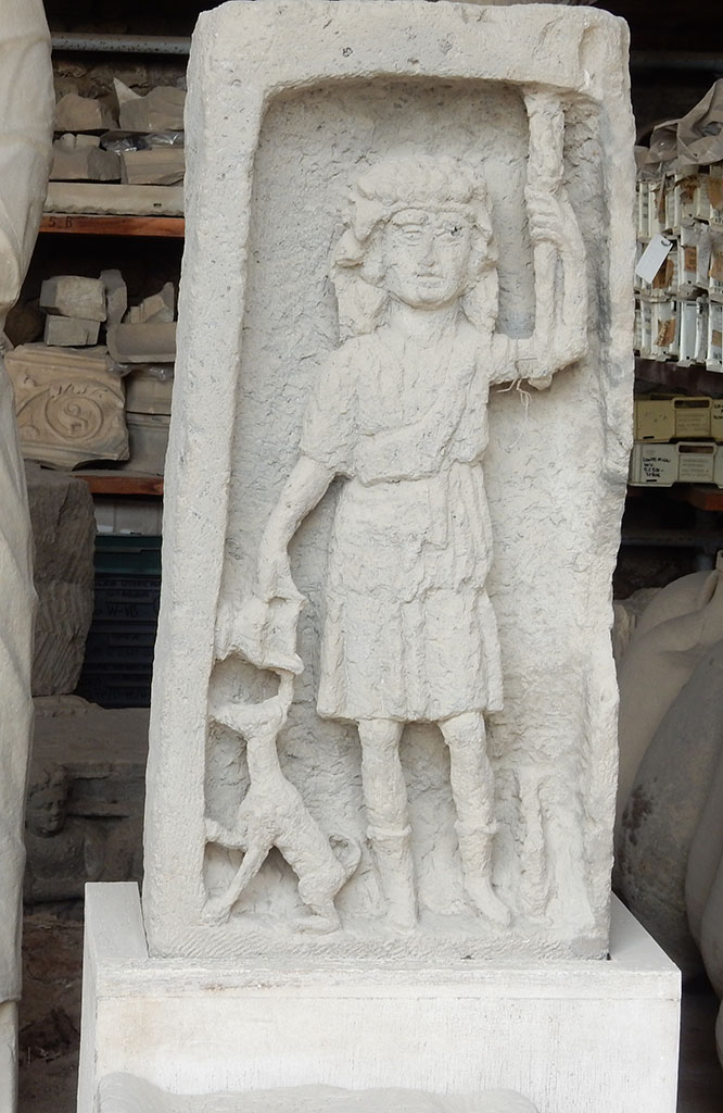 VII.7.29 Pompeii. May 2015. Bacchus stele in storage. Photo courtesy of Buzz Ferebee.
According to Kuivalainen, this a Bacchic votive stele of the Samnite era. 
It depicts a front facing skinny youth with his weight on his right foot, pouring wine from a cantharus for an animal rather resembling a dog than a panther.
The middle part of the thyrsus is missing.
The width of the frame varies, being thinner on the right hand side. 
It is cut deep into a rectangular upright tufa block and the workmanship is rather coarse.
The excavation details are not known.
PAP inventory number 8660.
See Kuivalainen, I., 2021. The Portrayal of Pompeian Bacchus. Commentationes Humanarum Litterarum 140. Helsinki: Finnish Society of Sciences and Letters, p. 207-8, H2.
