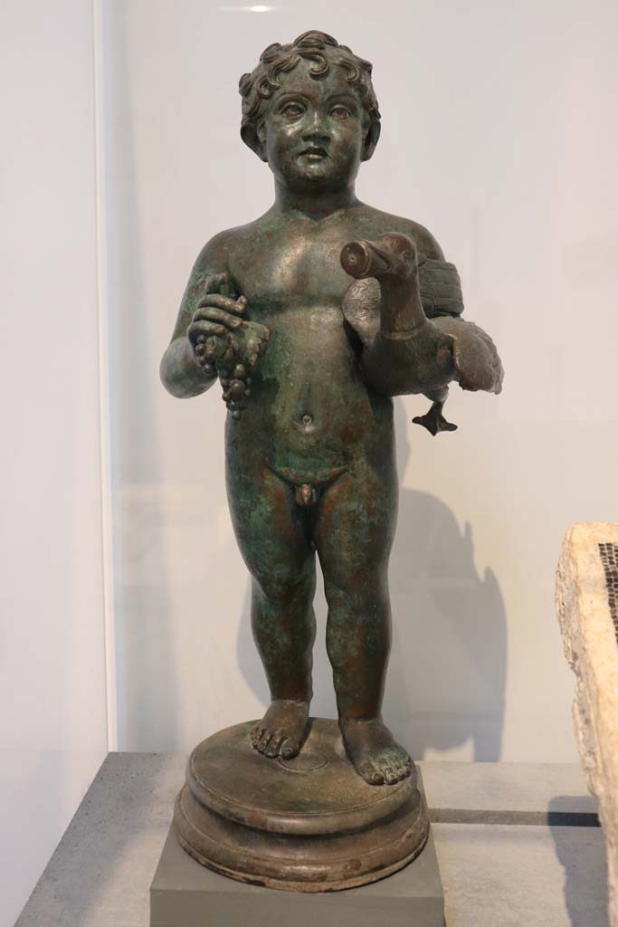 VIII.1.4 Pompeii. February 2021. One of the two bronze statuettes, (as described on P.107 above). 
Bronze statuette of young boy holding a goose and a bunch of grapes, from the peristyle of VI.15.1 (the House of Vettii).   
Photo courtesy of Fabien Bièvre-Perrin (CC BY-NC-SA).
