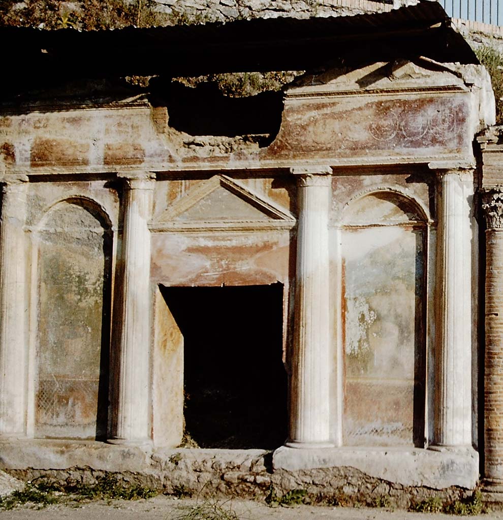 Pompeii Porta Nocera. 1959. Tomb 19ES. Richly decorated front of tomb with four columns, two top pediments, a lunette and stucco bordered panels. 
The front is richly decorated with garden paintings showing fences, marble crater fountains and shrubbery at the top of which are birds. 
Above the columns and below the top pediments are alternating and connected spirals of clovers and palmettes on a red background.
See D’Ambrosio, A. and De Caro, S., 1983. Un Impegno per Pompei: Fotopiano e documentazione della Necropoli di Porta Nocera. Milano: Touring Club Italiano. 19ES.
Detail from photo by Stanley A. Jashemski.
Source: The Wilhelmina and Stanley A. Jashemski archive in the University of Maryland Library, Special Collections (See collection page) and made available under the Creative Commons Attribution-Non-Commercial License v.4. See Licence and use details.
J59f0365
