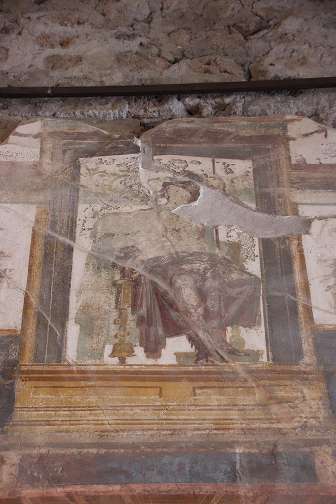 VI.15.1 Pompeii. October 2024. 
Upper south wall, painted seated central figure from above central panel. Photo courtesy of Klaus Heese.
In the upper centre of the south wall, damaged, a figure with left shoulder destroyed, and fractures throughout the painting. 
Kuivalainen describes – 
“Inside an aedicula on a white background, a youth sits on a throne with his legs on a footstool: he leans with his left hand on a long thyrsus and wears high boots and a red cloak on his hips: the yellowish legs of the throne are turned. 
Kuivalainen comments –
“The whole south wall is thematically Bacchic, and this young half-naked Bacchus is depicted among other gods in the architecture of the upper zone.”
See Kuivalainen, I., 2021. The Portrayal of Pompeian Bacchus. Commentationes Humanarum Litterarum 140. Helsinki: Finnish Society of Sciences and Letters, (p.100-1, B11).
